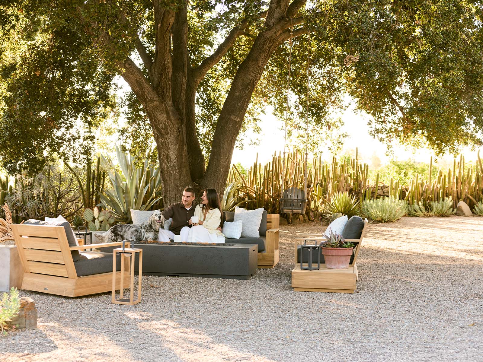 Sonora outdoor patio furniture set under a large tree with a couple sitting on a dark gray sectional sofa, a dog on a cushion, and matching teak lounge chairs and side tables on a gravel courtyard.