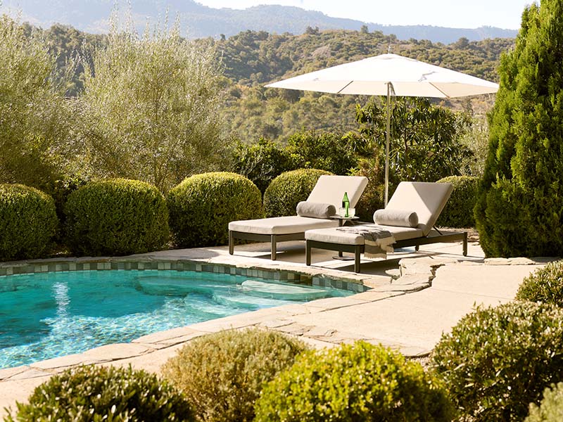 Outdoor patio scene with two beige lounge chairs under a large white umbrella beside a swimming pool, surrounded by manicured green shrubs and trees with hills in the background.