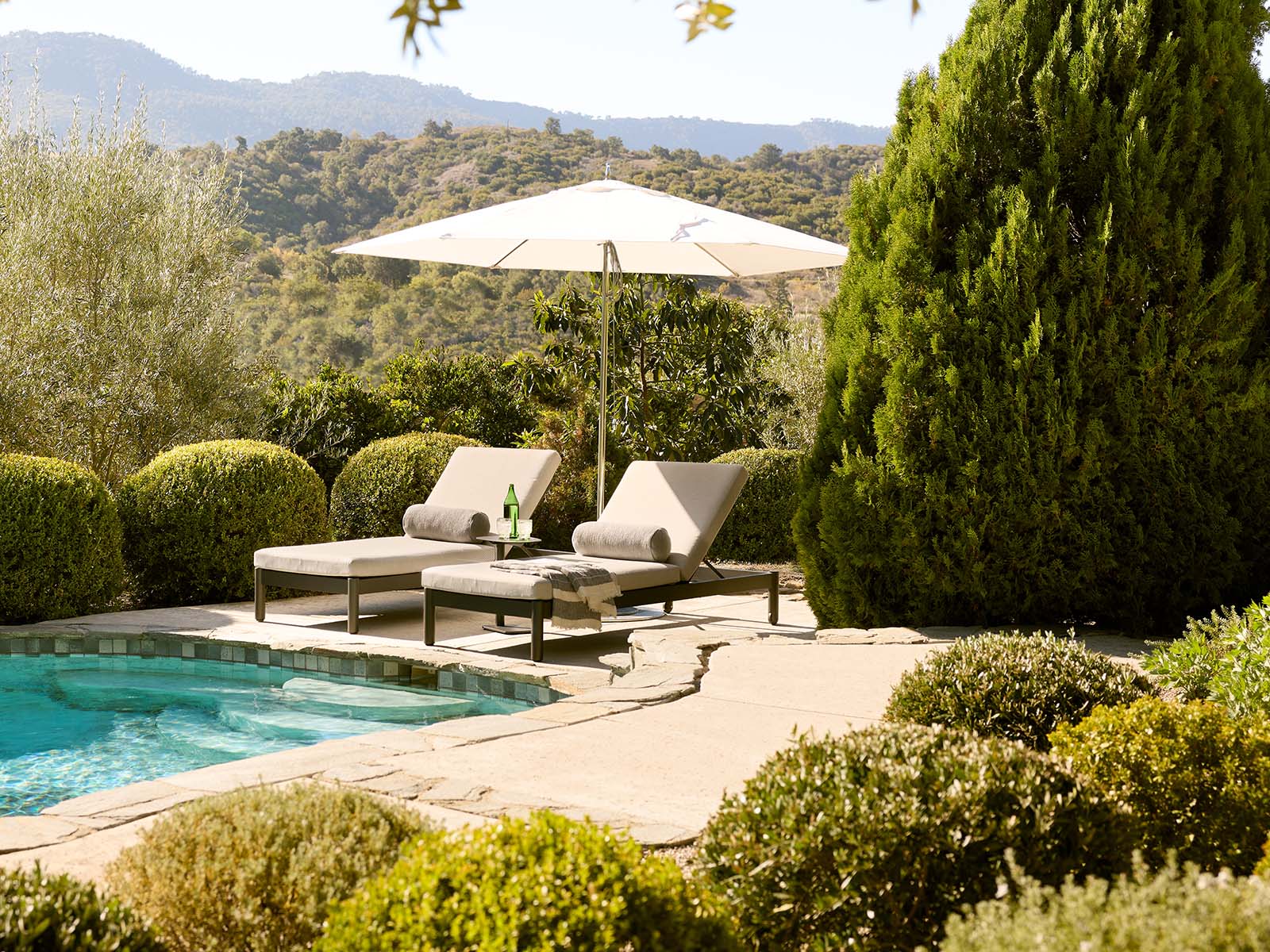 Outdoor patio scene with two beige lounge chairs under a large white umbrella beside a swimming pool, surrounded by manicured green shrubs and trees with hills in the background.