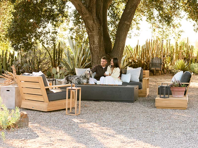 Sonora outdoor patio furniture set under a large tree with a couple sitting on a dark gray sectional sofa, a dog on a cushion, and matching teak lounge chairs and side tables on a gravel courtyard.